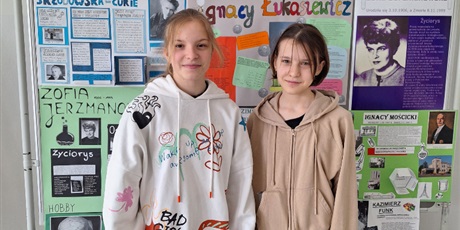 Powiększ grafikę: Uczennice Ala i Maja stoją na tle gazetki o wielkich polskich chemikach